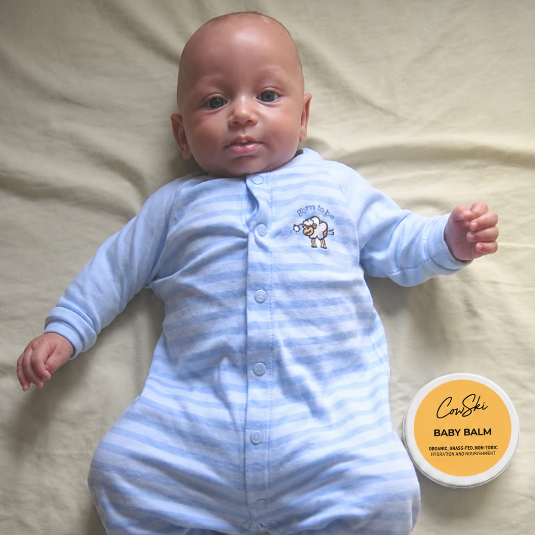Baby in a blue onesie lying on a beige surface next to a container of baby balm.