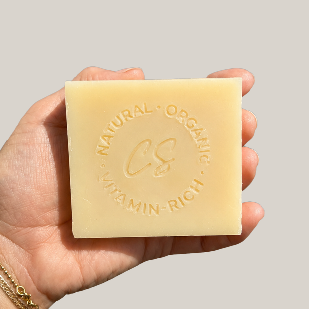 A hand holding a square, pale yellow bar of soap with embossed text reading 'Natural Organic Vitamin Rich CS' against a solid light gray background.