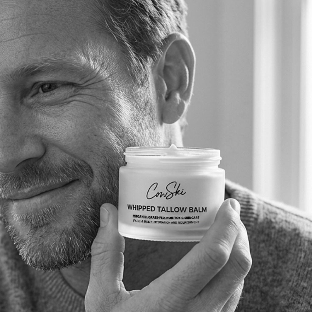 A man holds a white jar of CowSti Whipped Tallow Balm close to his face in a softly lit indoor setting. The image is in black and white, showing the man's smiling face, hand, and the product jar with clear text.